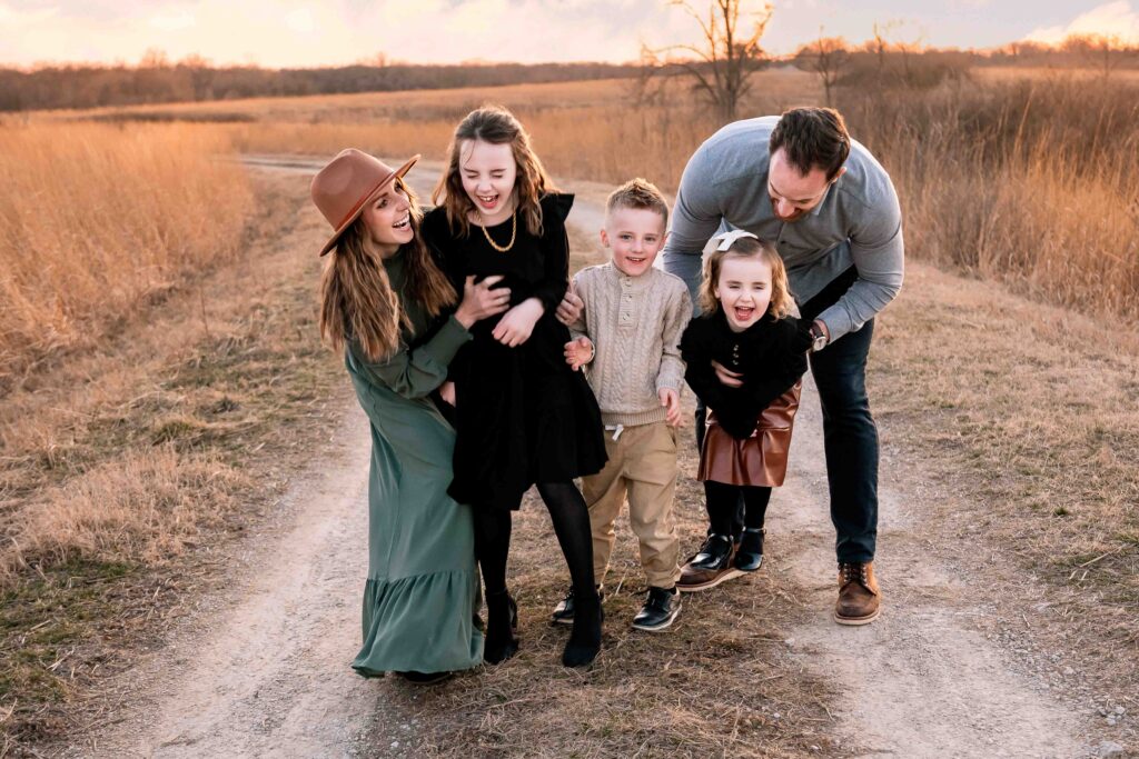 Happy family of five laughing together on a rustic dirt path surrounded by golden fields at sunset. Parents and three stylishly dressed children share a joyful moment outdoors.