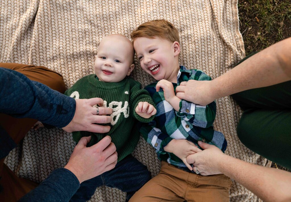 Joyful moment at the St. Louis Botanical Garden as two young brothers lie on a cozy knit blanket, giggling while being playfully tickled by their parents. The baby smiles in a green sweater while his older brother laughs in a green plaid shirt—capturing pure sibling happiness in this relaxed, family-centered setting.