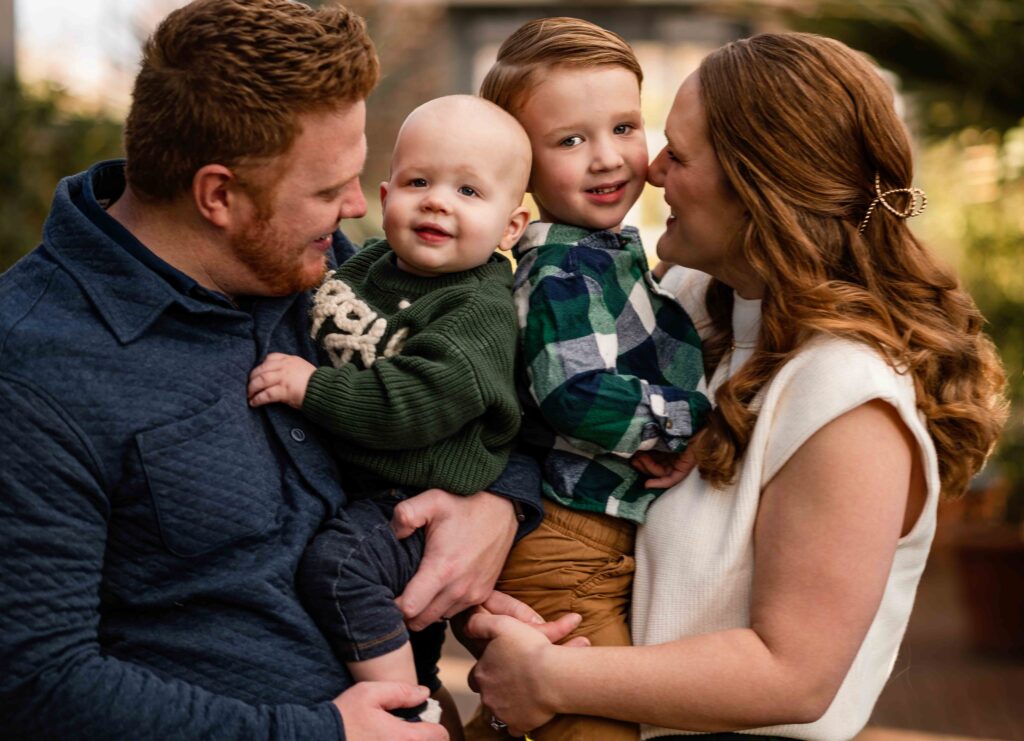 Candid family portrait at the St. Louis Botanical Garden featuring parents holding their two young sons. The baby smiles sweetly in a cozy green sweater while the older brother snuggles close, surrounded by natural light and lush indoor greenery—capturing warmth, connection, and joy in this beautiful St. Louis setting.
