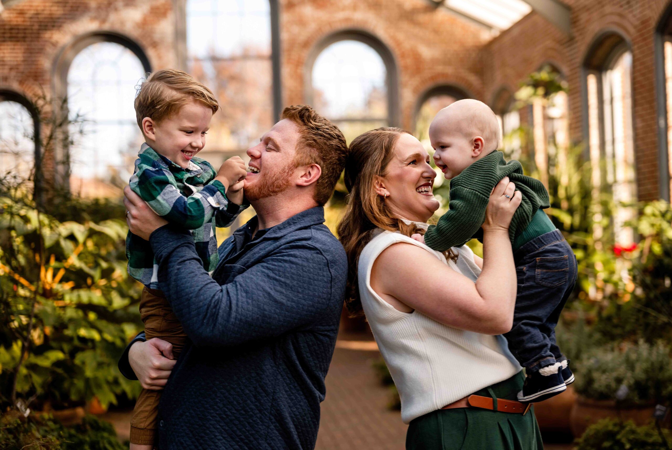 Joyful family of four shares a playful moment inside the historic brick conservatory at the St. Louis Botanical Garden. Dad lifts his laughing toddler while mom lifts their smiling baby, surrounded by lush greenery and soft natural light—perfect for a candid, heartwarming family session in St. Louis.
