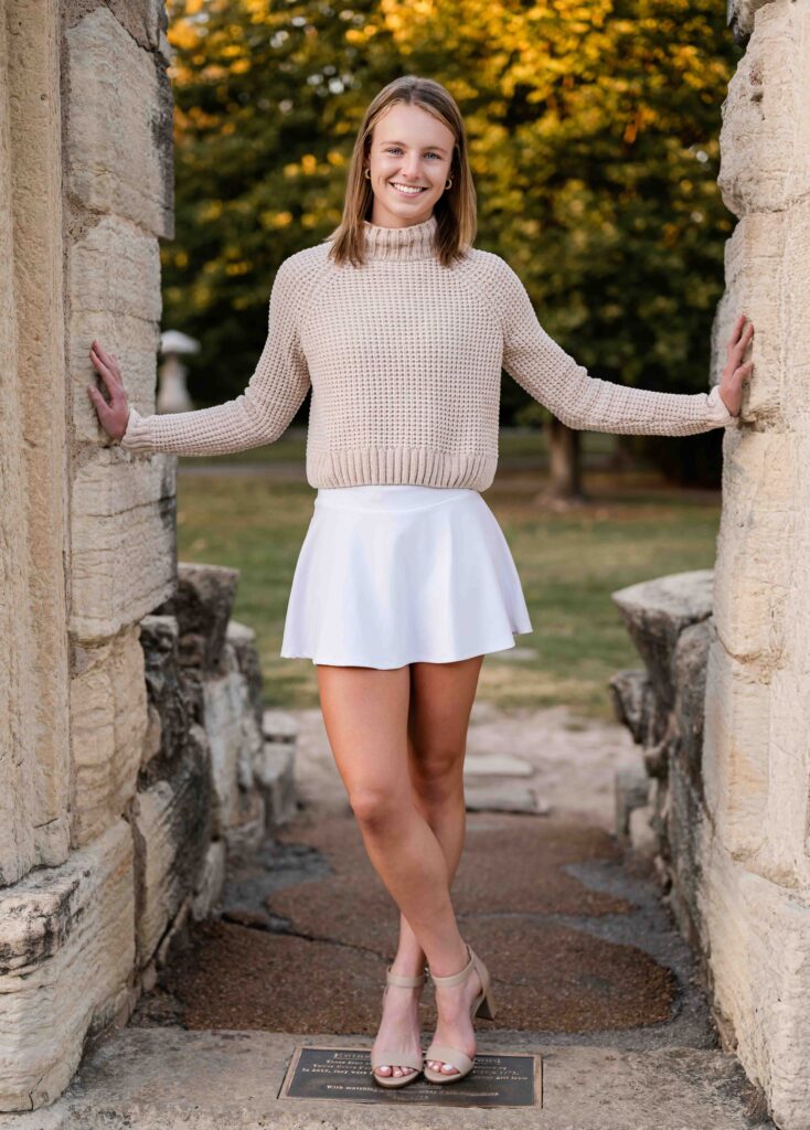 Teen girl in a white sweater and skirt leaning against a stone structure surrounded by fall foliage in Tower Grove Park.