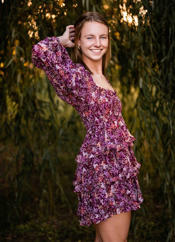 High school senior posing at willow tree in a maroon dress with one hand raised behind her head at Tower Grove Park, St. Louis.