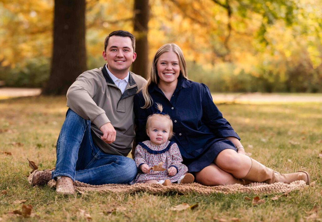 Family sitting together on a blanket in a sunlit park, smiling and cuddling during a fall photo session in St Louis, Missouri.