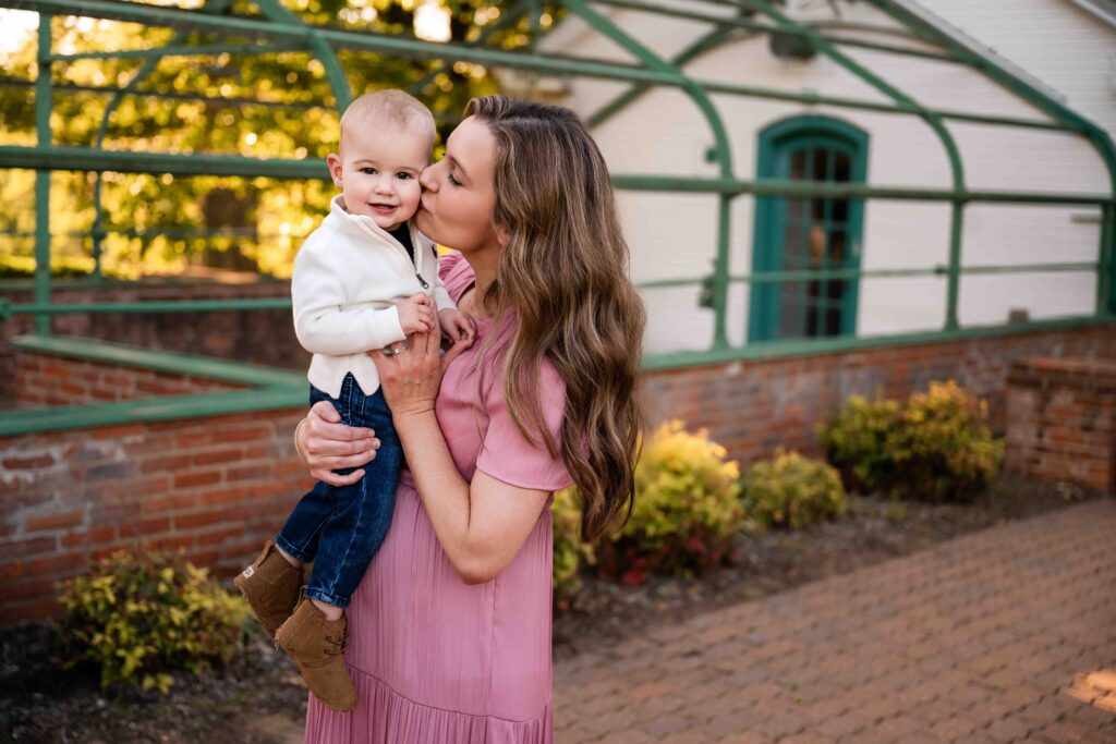 Mother kissing baby on the cheek during outdoor family photo session in St. Louis, natural light photography against brick wall and garden greenhouse background