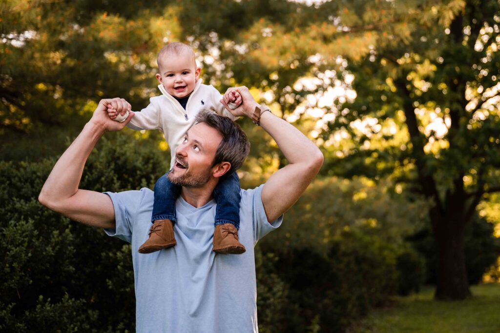 Father holding baby on his shoulders during outdoor family photo session in St. Louis, golden hour natural light photography with green trees in background