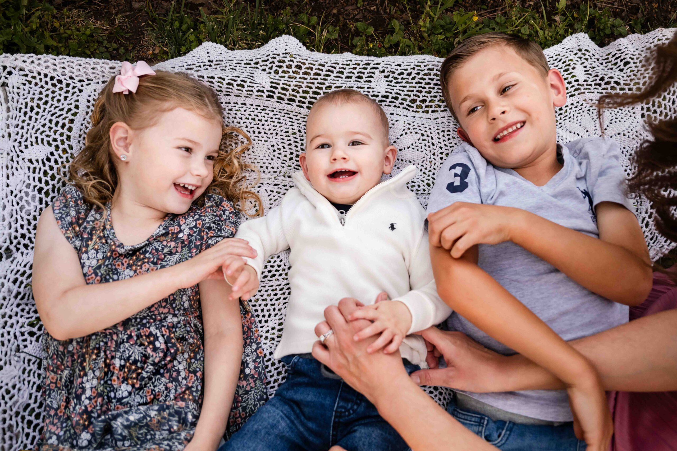 Three siblings lying on a blanket laughing during outdoor family photo session in St. Louis, natural light photography of brother and sisters with baby