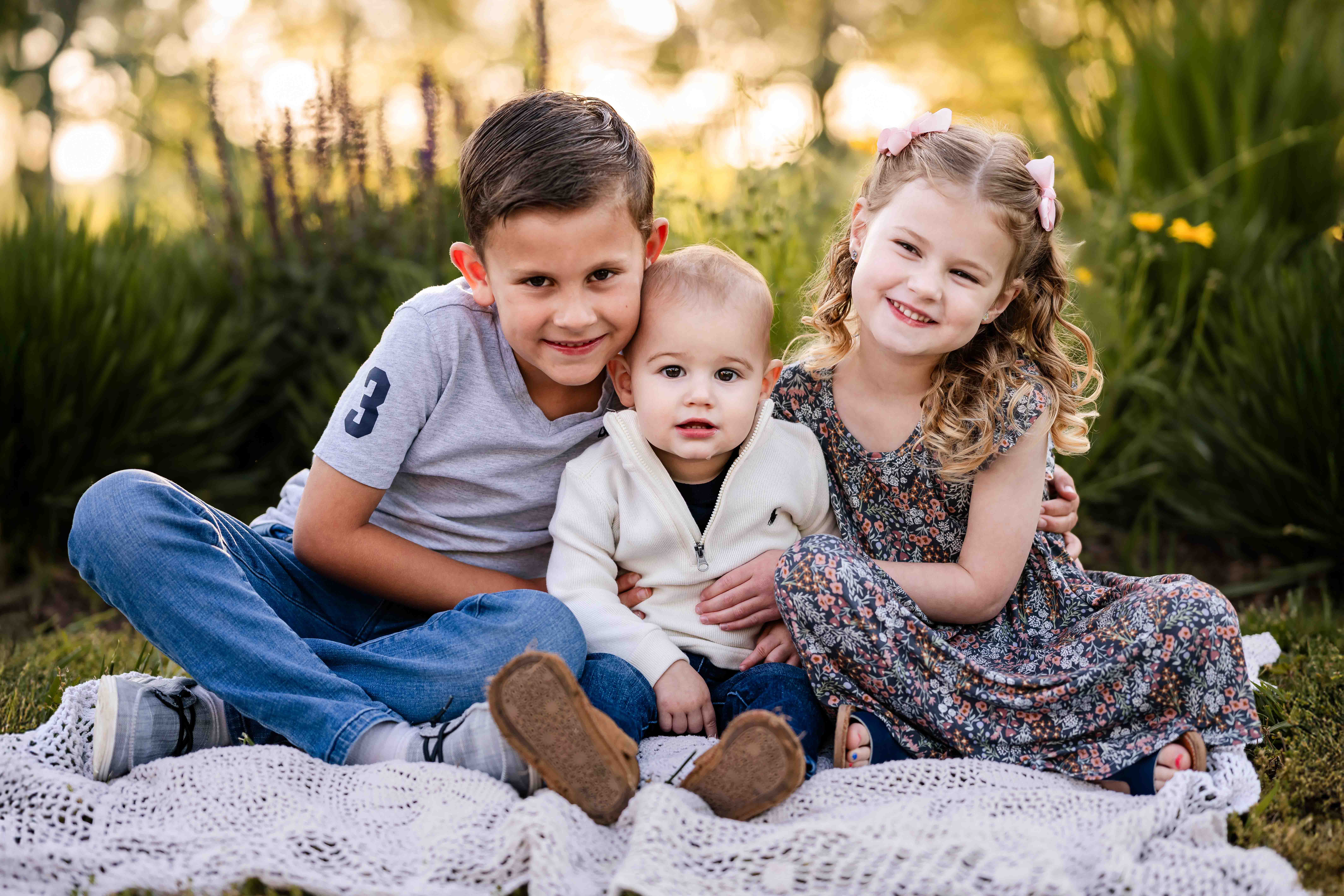 Three siblings sitting together on a blanket during golden hour outdoor family photo session in St. Louis, natural light sibling portrait with garden background