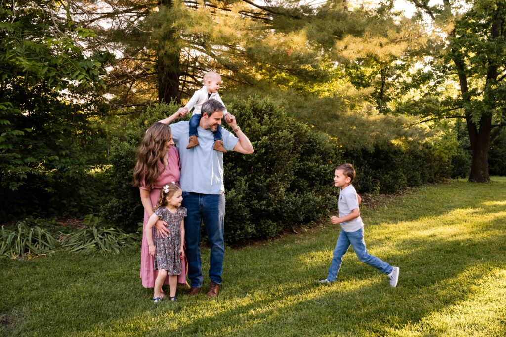 Family of five during outdoor golden hour photo session in St. Louis, parents with three kids including baby on dad's shoulders and boy running through grass, natural light family photography