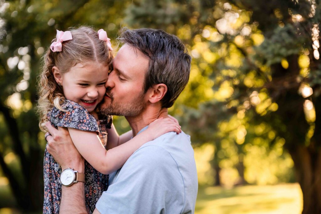 Father kissing little girl on the cheek during golden hour outdoor family photo session in St. Louis, natural light dad and daughter portrait with bokeh tree background