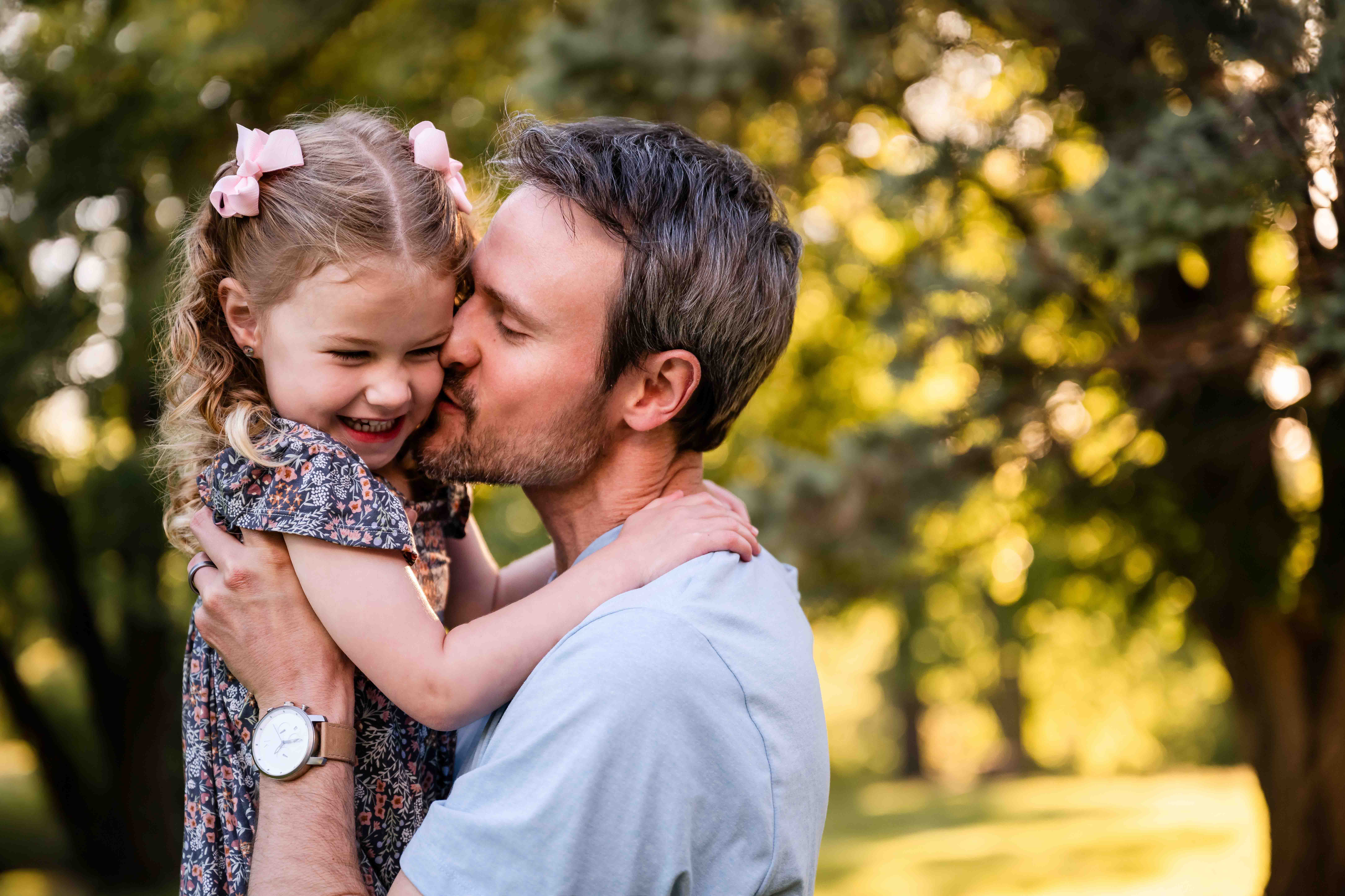 Father kissing little girl on the cheek during golden hour outdoor family photo session in St. Louis, natural light dad and daughter portrait with bokeh tree background