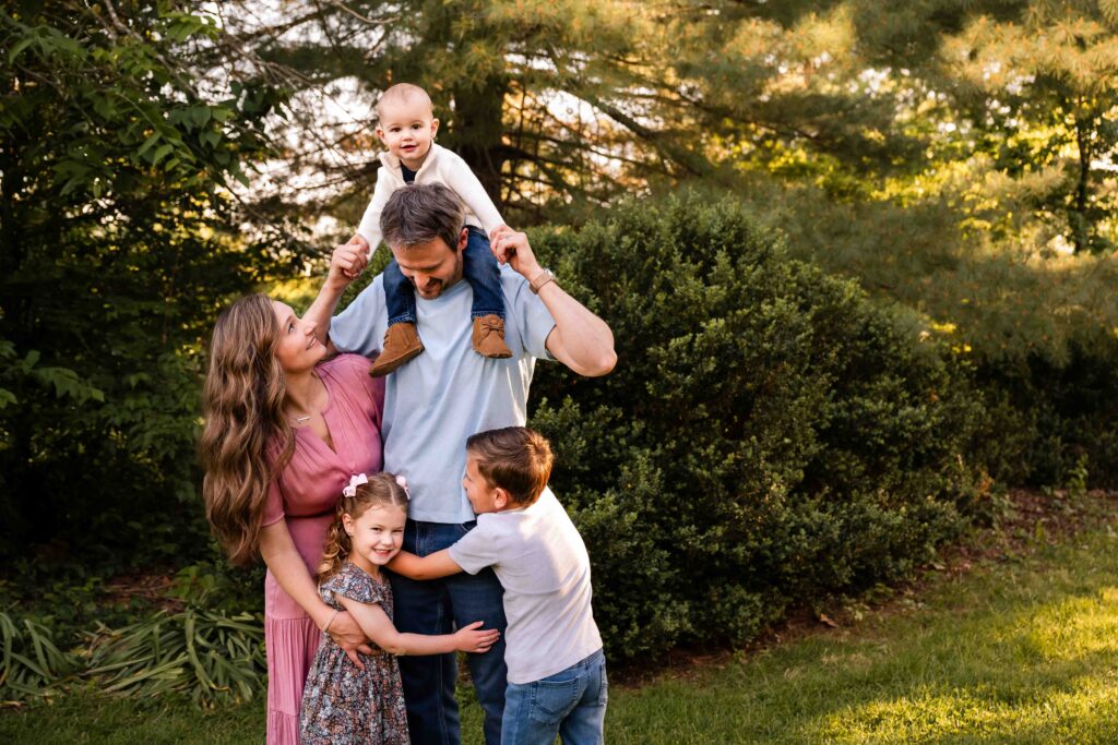 Family of five together during golden hour outdoor family photo session in St. Louis, parents and three kids with baby on dad's shoulders, natural light family portrait in green garden setting